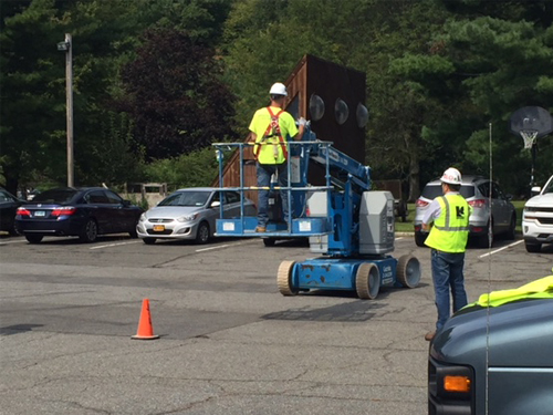 Two construction workers in a parking lot, One on the ground and one in an aerial lift.