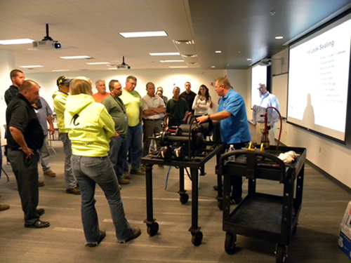 A large group of construction workers huddled in front of man and a cart who is doing a training.