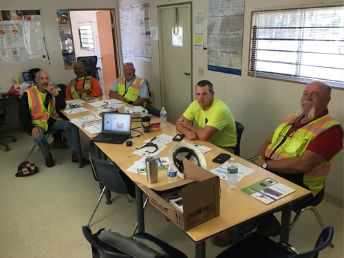 Five construction workers sitting around table in a trailer with computer and papers.