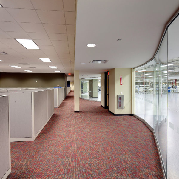 Office hallway with cubicles to the left and glass wall looking into manufacturing space to the right